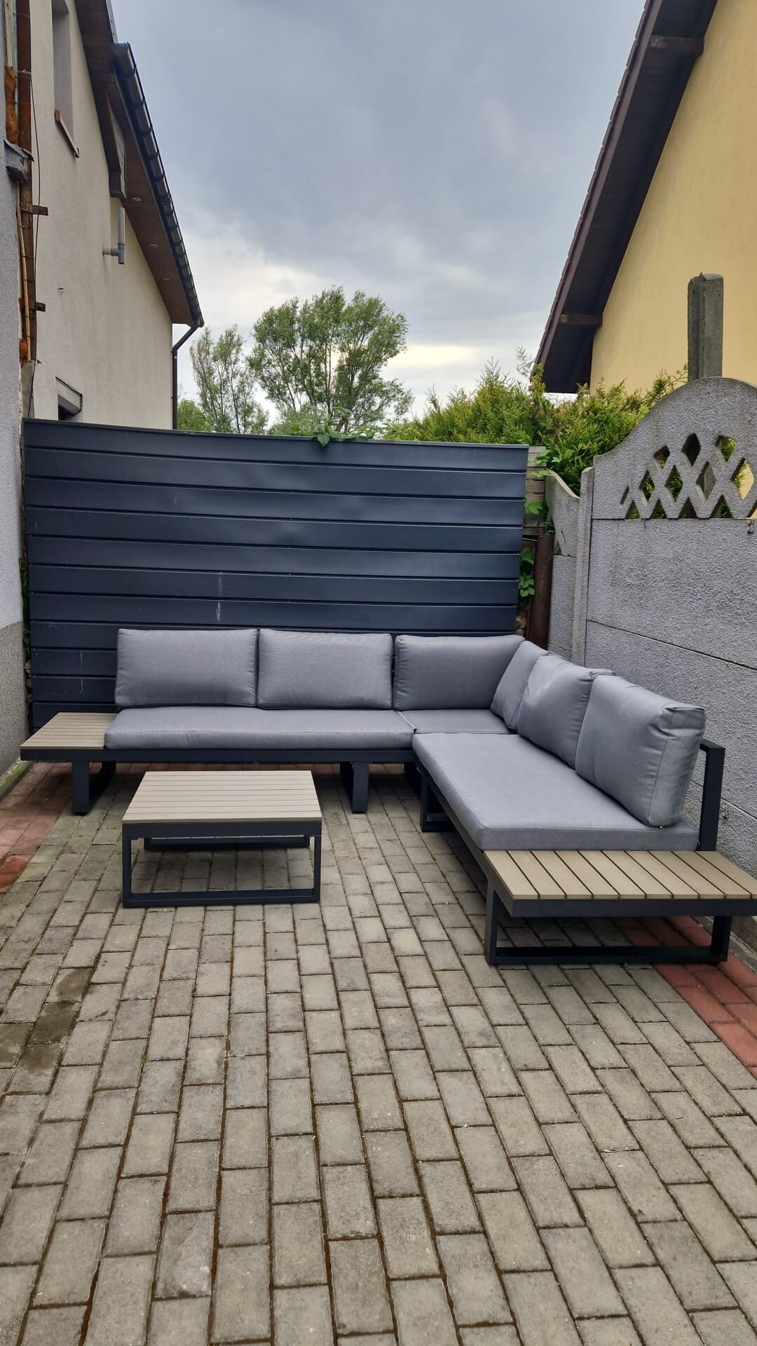 An outdoor seating area is presented, featuring a sectional sofa with gray cushions arranged around a small, rectangular coffee table. The seating is positioned on a paved patio in a cozy garden space, framed by a wooden privacy wall and lush greenery.