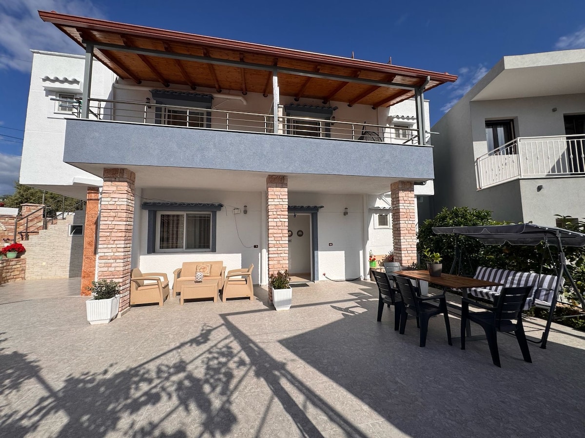 A well-structured two-story building features a welcoming outdoor patio with a dining table and seating arrangements. Natural light creates defined shadows across the surface. The upper balcony, supported by brick columns, offers additional space for relaxation.