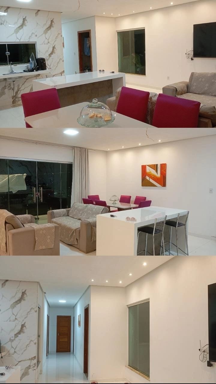 A spacious living area features modern furniture with a mix of cozy couches and dining chairs in vibrant pink. A sleek countertop separates the kitchen from the living space, while large windows allow natural light to fill the room. Minimalist artwork decorates the walls.