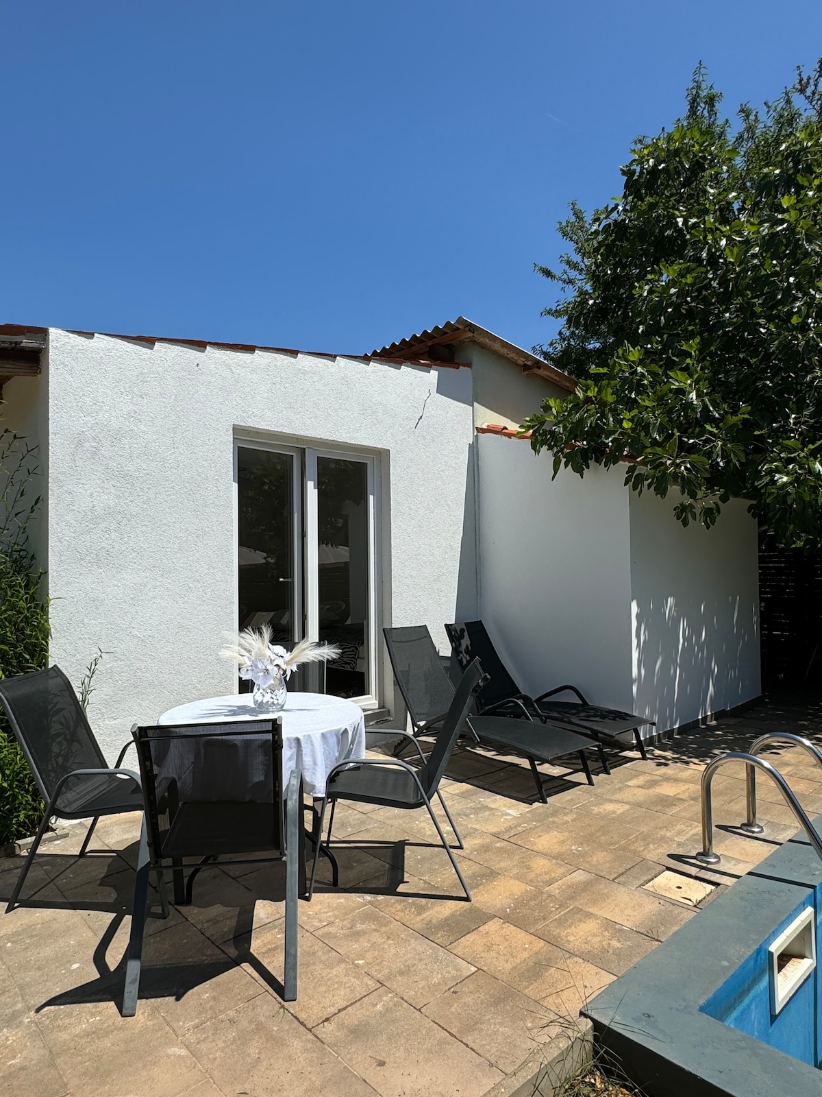 A spacious outdoor area is visible, featuring a round table with a white tablecloth and four chairs. Two lounge chairs are positioned nearby, all placed on a tiled surface. Natural greenery surrounds the space, and a portion of a swimming pool is seen in the corner.