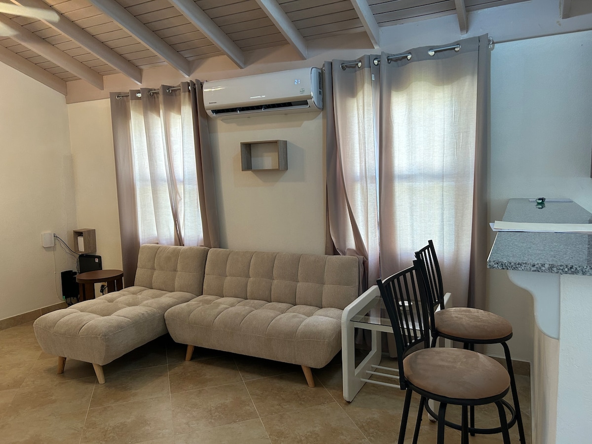 A light-filled living area is presented, featuring a comfortable sectional sofa with a neutral fabric. Adjacent to it, a small round dining table with two bar stools is visible. Soft curtains frame the windows, while a mini-split air conditioning unit is mounted on the wall.