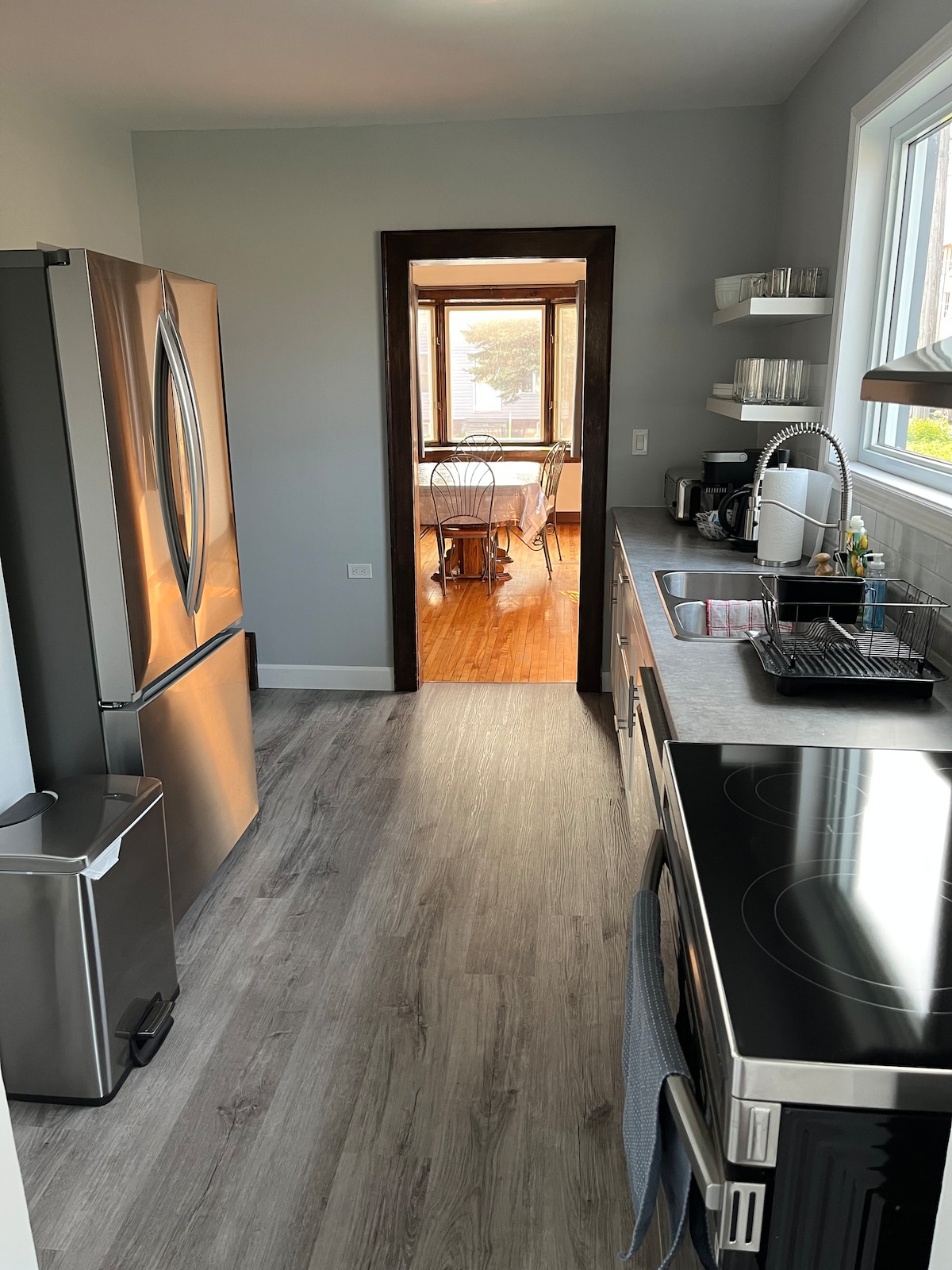 An updated kitchen features stainless steel appliances, including a refrigerator and stove with an oven. A doorway leads to a dining area, where natural light enhances the wooden floors and simple table setting. Countertop space is organized, with a sink and kitchen utensils visible.