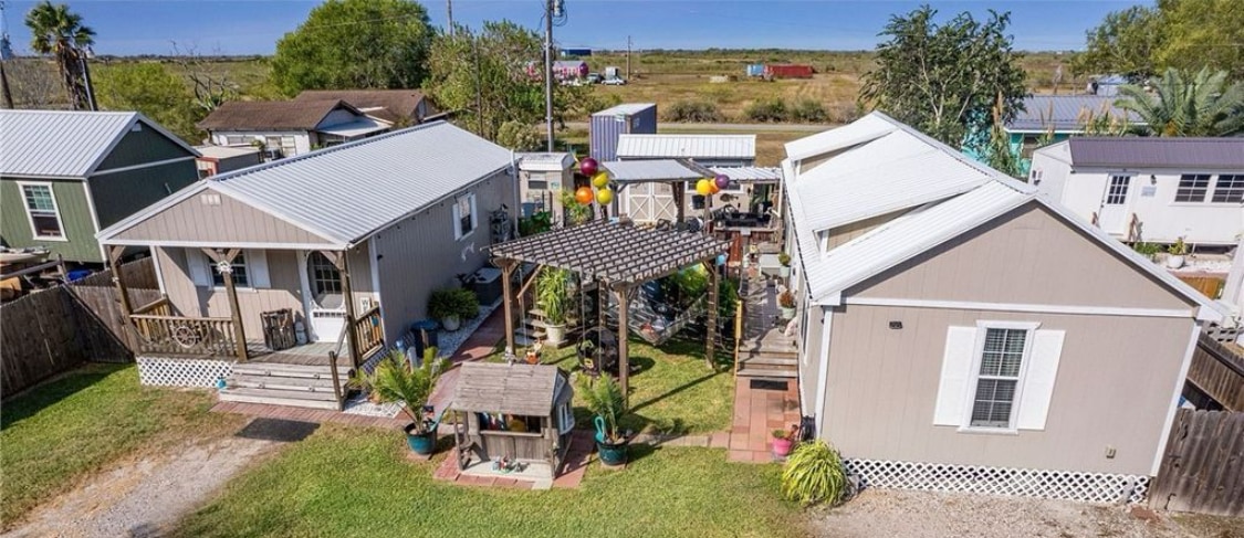 An aerial view captures three homes surrounded by green grass and a gravel pathway. Outdoor seating is visible under a wooden pergola draped with colorful lanterns. Lush plants and patio furniture enhance the outdoor space, creating a welcoming area for relaxation.