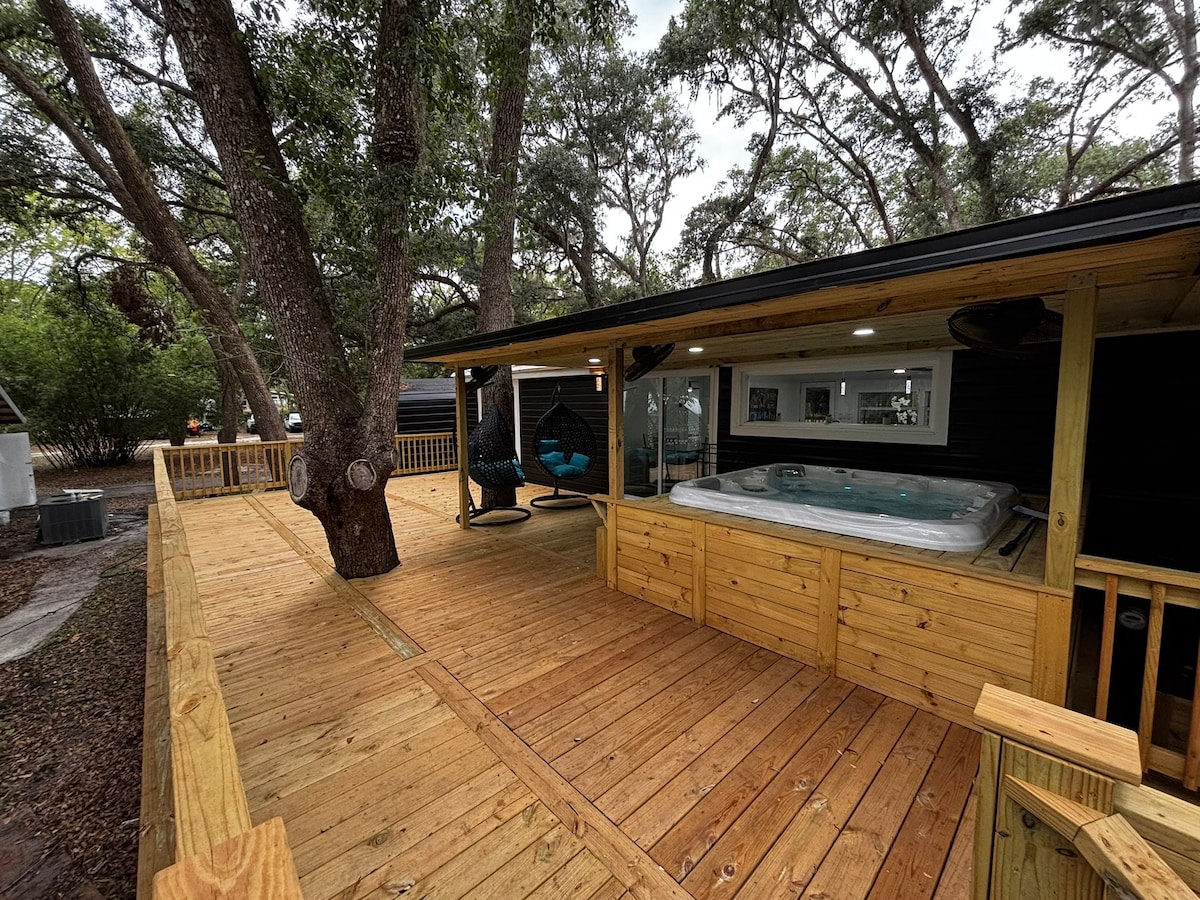 An expansive deck made of warm wood provides ample space for relaxation, featuring a jacuzzi positioned under the shade of a large tree. Comfortable hanging chairs are visible, ideal for enjoying the outdoor setting. The modern home can be seen in the background, enhancing the serene atmosphere.