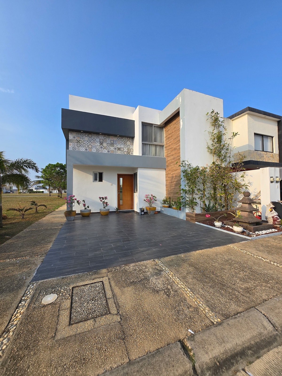 A modern two-story house is depicted, featuring a mix of white and wooden exterior elements. The entrance is framed by colorful flower pots. A well-maintained walkway leads to the door, while adjacent buildings and greenery are visible in the background under a clear blue sky.