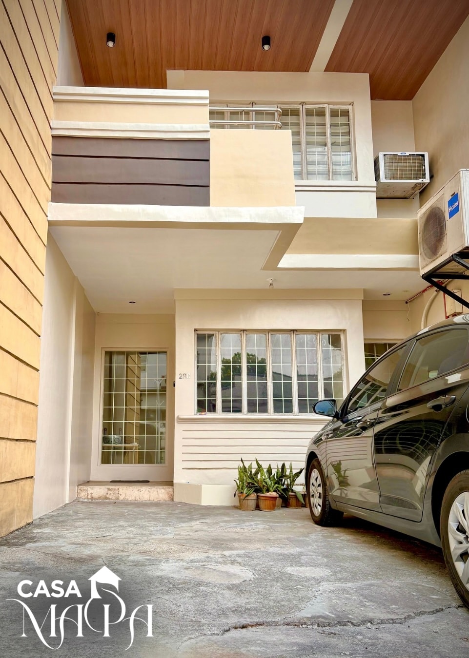 A modern entrance showcases a well-maintained façade with a combination of light colors. Large windows provide natural light, and potted plants add a touch of greenery. A parked car is visible in front of the entrance, which is set against a textured stone surface.