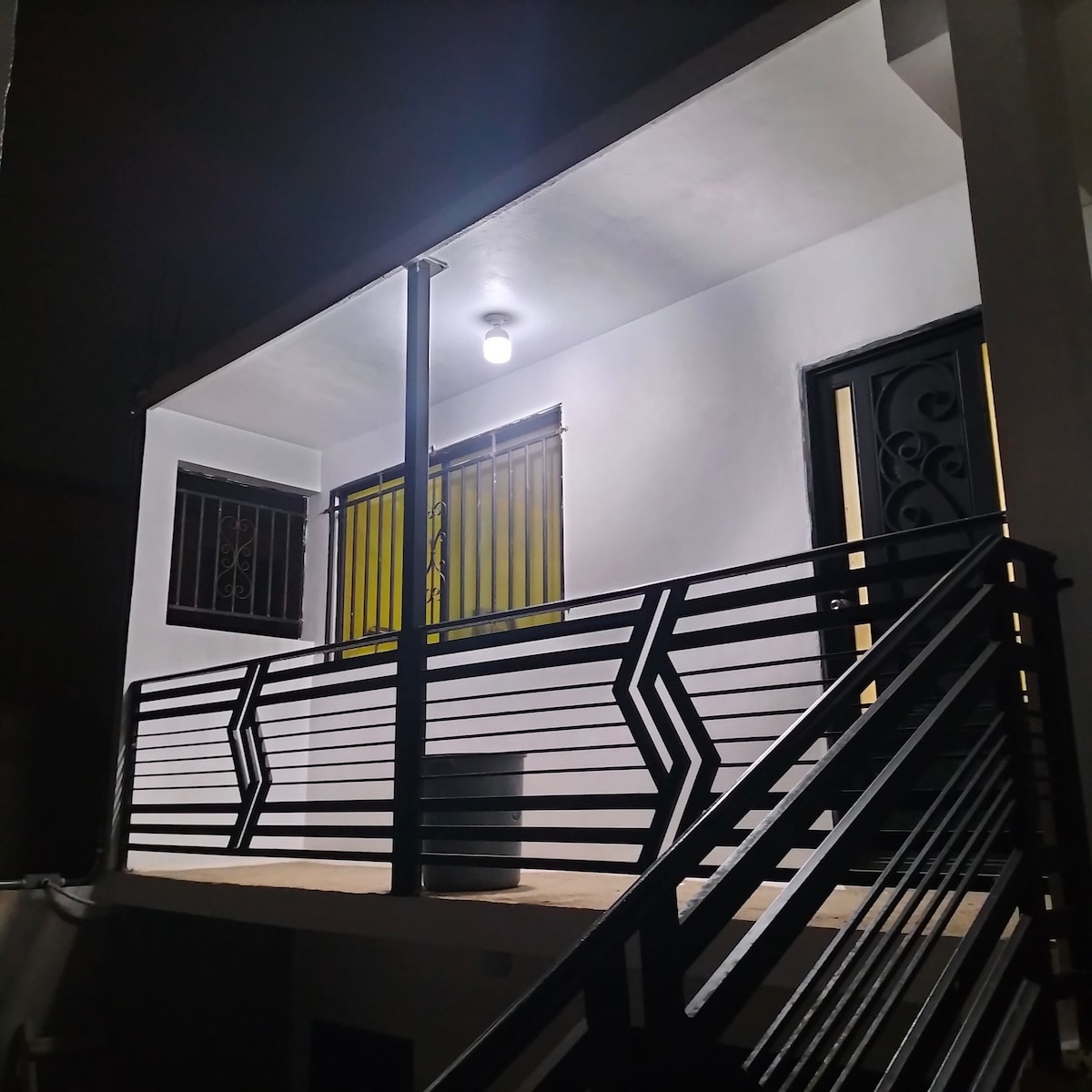 The exterior of a welcoming home is presented, featuring a simple balcony with modern railing design. A bright light fixture illuminates the entryway, while yellow curtains peek through the window nearby, providing a hint of warmth amidst the clean white facade.