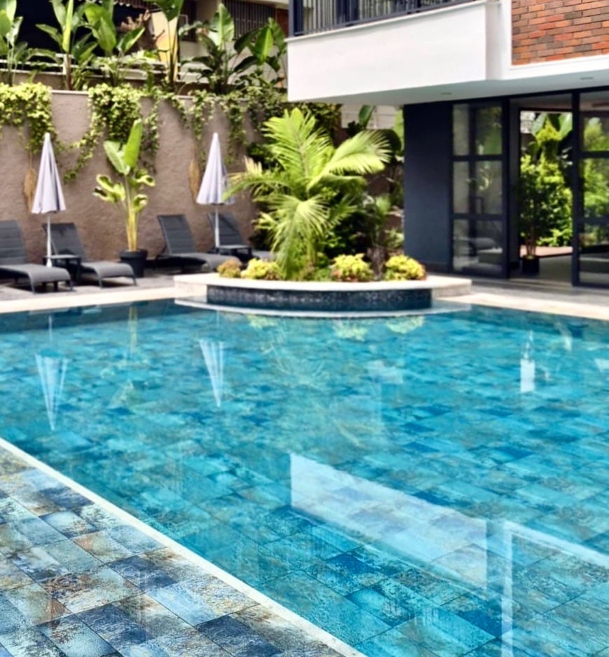A tranquil outdoor pool is surrounded by lush greenery and elegant lounge chairs. Palm trees provide a tropical touch, while a circular garden feature adds to the serene atmosphere. Sun umbrellas line the poolside, offering shaded areas for relaxation.