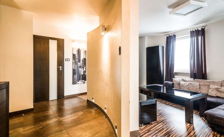A spacious living area features a sectional sofa and a dark coffee table positioned on patterned wood flooring. Dark curtains frame a window, allowing natural light to brighten the space. The curved wall leads to a hallway with modern artwork, adding an elegant touch.
