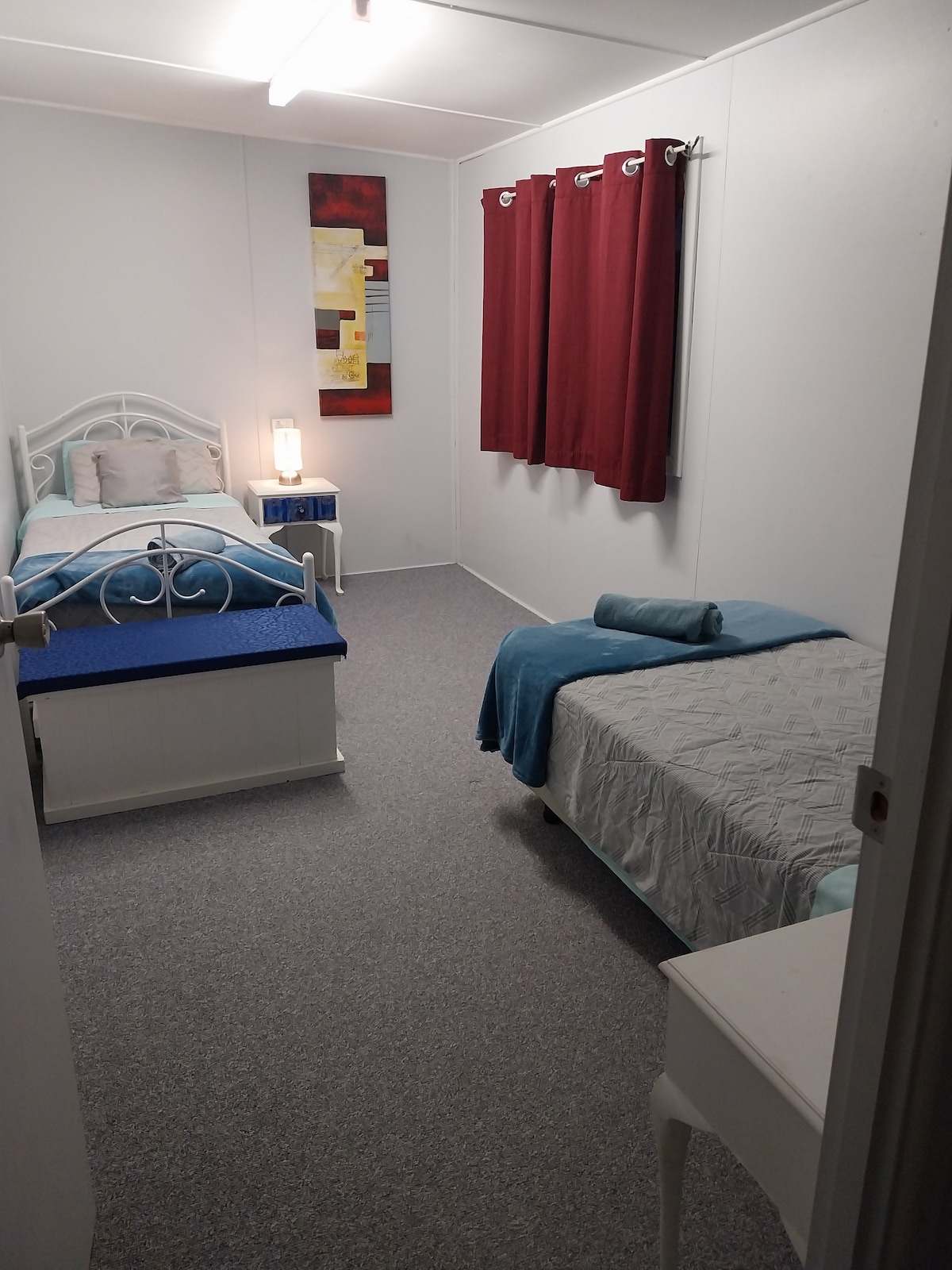 A well-lit bedroom is shown, featuring two single beds with varying bedding colors. A light blue blanket is neatly arranged on one bed, while a decorative throw pillow is placed on the other. A small bedside table with a lamp and a piece of wall art are visible. Dark red curtains frame a window, enhancing the room's cozy appearance.