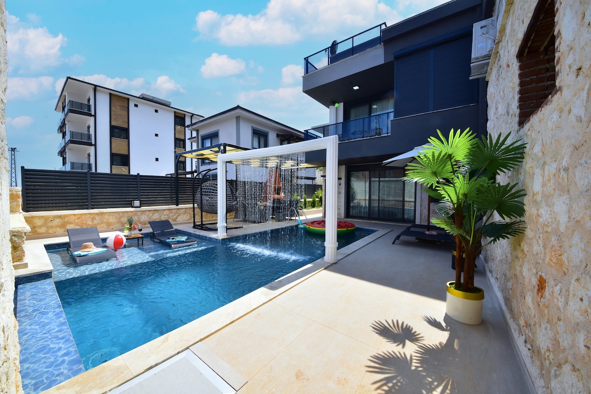 A spacious outdoor area features a glimmering swimming pool surrounded by lounge chairs and colorful floaties. An inviting leisure space is enhanced by a covered seating area. Lush greenery is present in a potted plant, contributing to the serene environment. Modern structures form the backdrop.