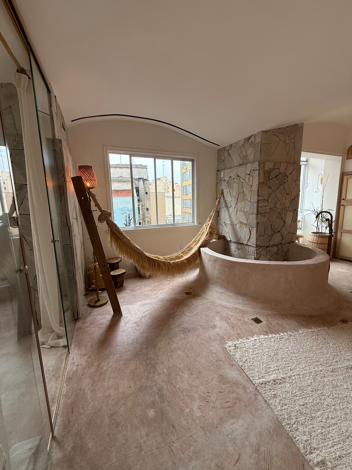 A spacious bathroom features a circular stone bathtub and a soft, textured rug on the floor. Natural light fills the room from a large window, while a hammock is gently suspended nearby. Glass panels separate the shower area, enhancing the open and airy feel.