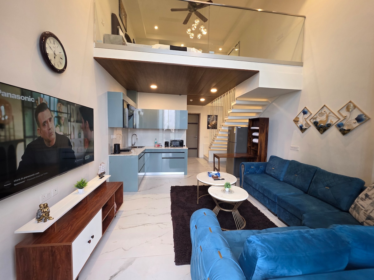 A spacious living area is featured, showcasing a plush blue sectional sofa and a stylish center table on a textured rug. A modern TV unit with a flat-screen TV is mounted on the wall. The open kitchen area is visible behind, highlighting sleek cabinetry and contemporary appliances.