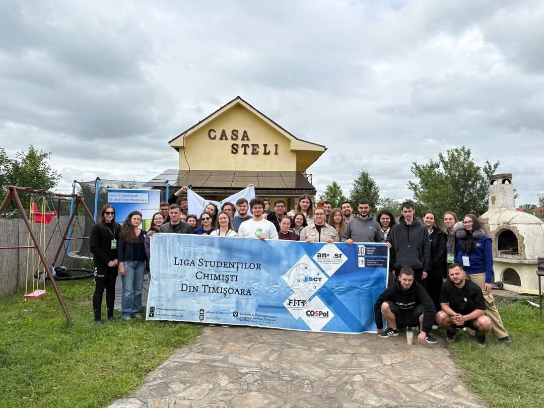 A group of individuals stands in front of the Casa Steli building, which features a prominent sign. Green grass and trees are visible in the background, alongside a swing set and a barbecue area. A large banner is held by the group.