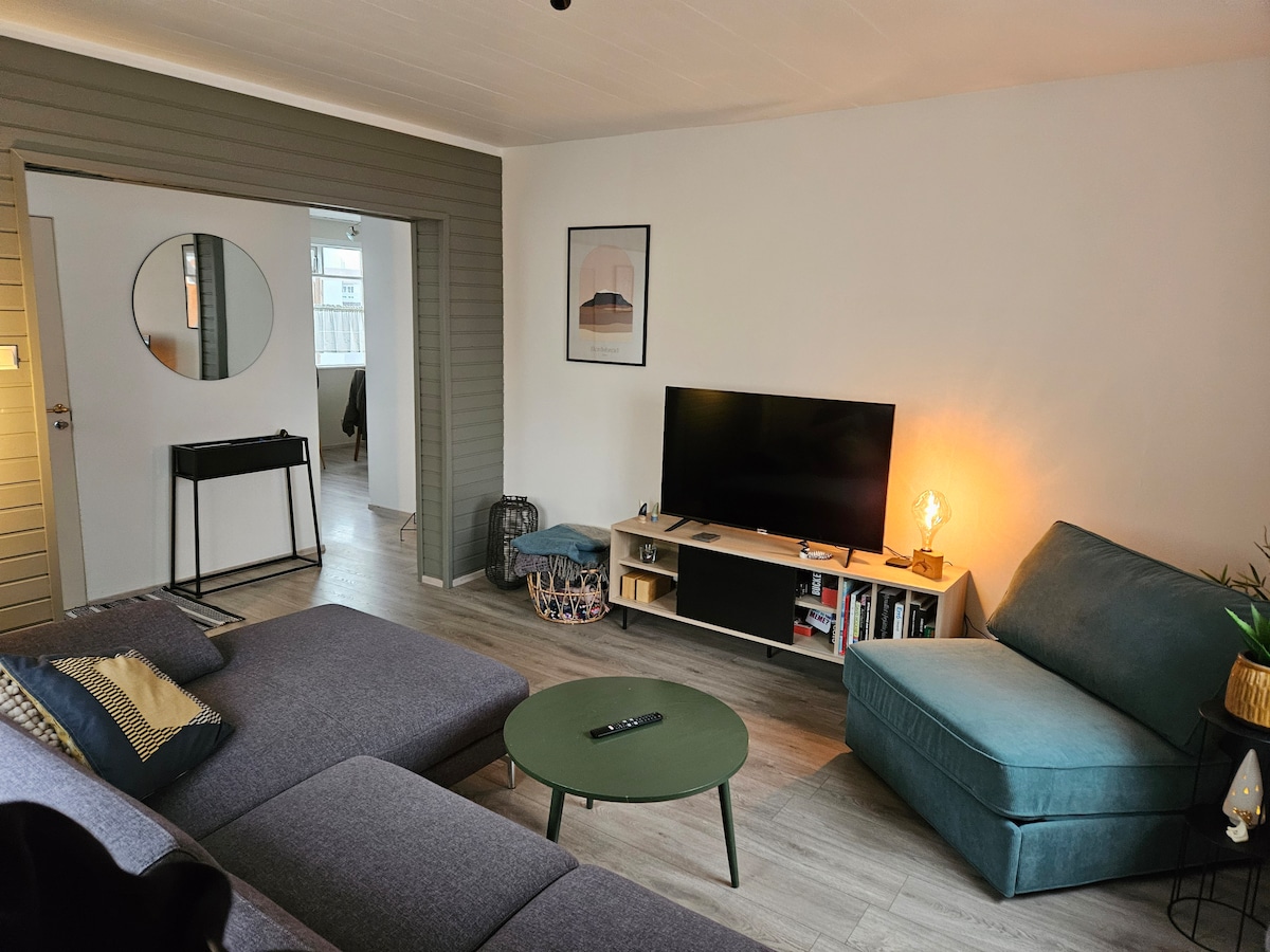 A living room area is presented with a cozy sectional sofa and a teal armchair. A low circular table with a green finish is centered between the seating. A TV unit with various decorative items and a warm lamp adds a soft glow to the space. A doorway leads to another room.