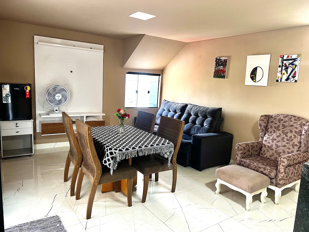 A spacious living area features a dark sofa and a patterned armchair, complemented by a wooden dining table surrounded by chairs. Natural light enters through a window, and decorative art pieces adorn the walls. A fridge and fan are visible in the background.