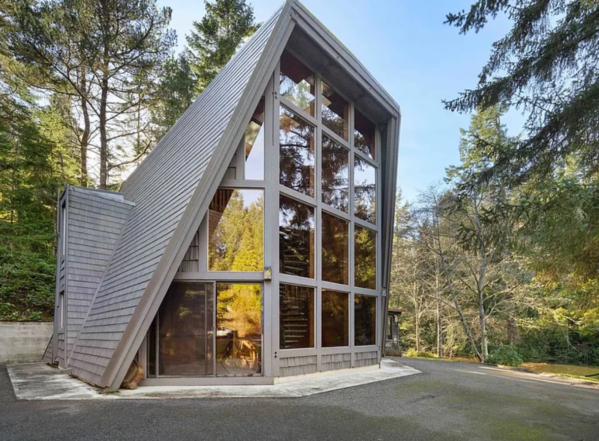The exterior of a modern glass house is showcased, featuring a distinctive A-frame design. Large windows reflect the surrounding trees and landscape, allowing natural light to illuminate the space. A paved driveway provides access to the entrance, which is framed by lush forest.