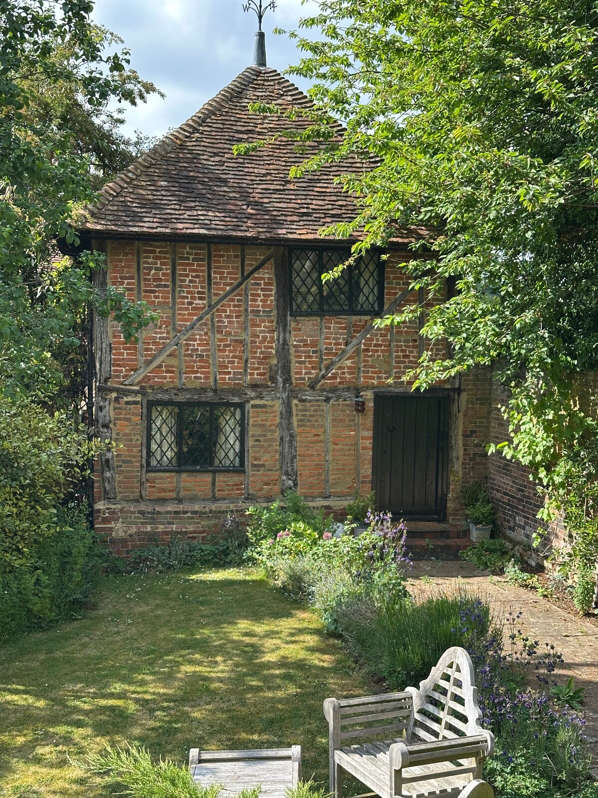 The exterior of the historic Dove Cote is showcased, featuring timber-framed construction with exposed brick. The entrance door is framed by lush greenery, while a small garden area is visible, populated with flowering plants and a wooden bench inviting relaxation.