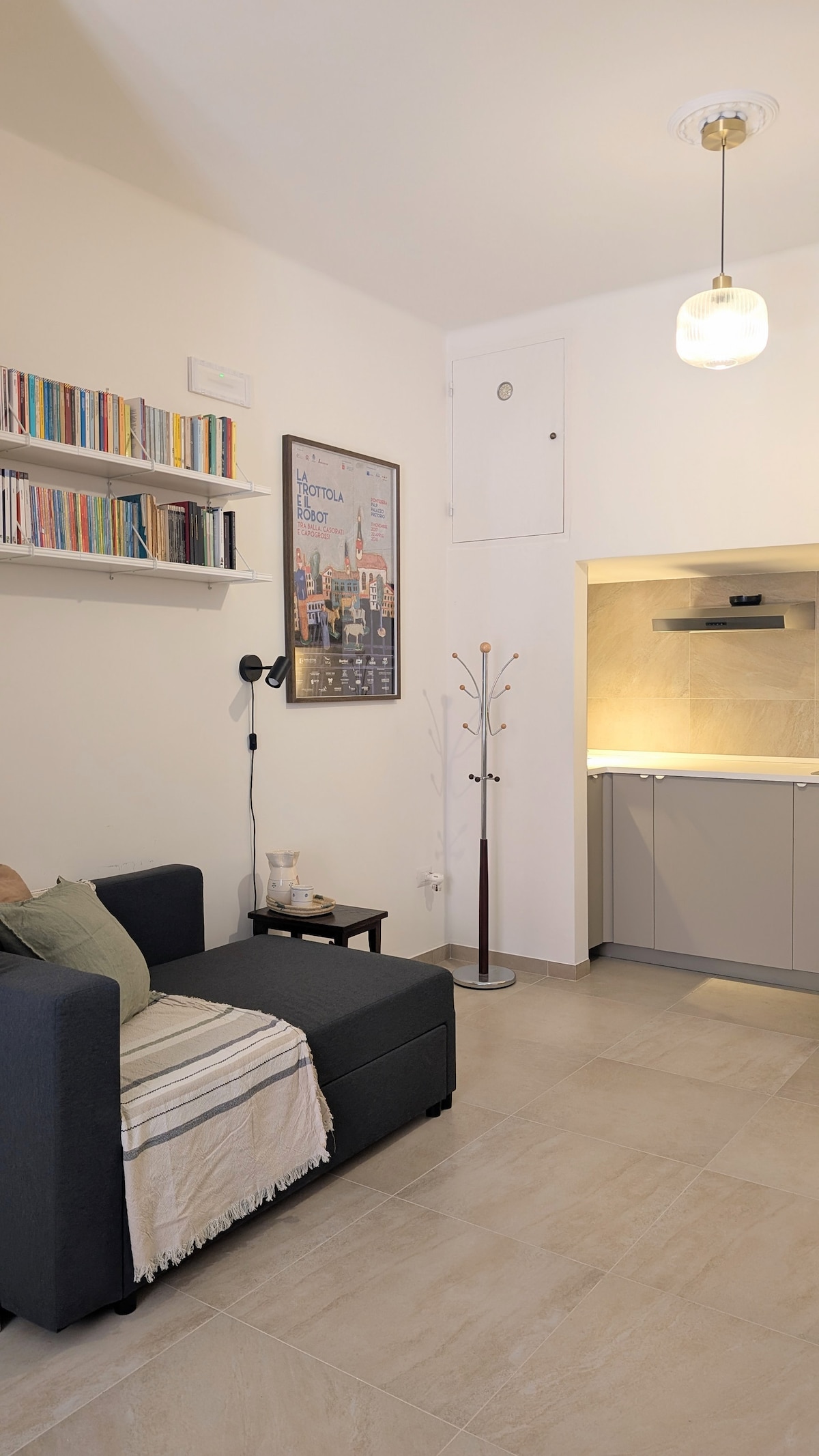 A compact living area features a dark sofa adorned with a light blanket. A wall-mounted shelf displays various books, while a framed city poster adds visual interest. A stylish lamp illuminates the space, and the corner showcases a modern coat rack, complementing the kitchen area in the background.