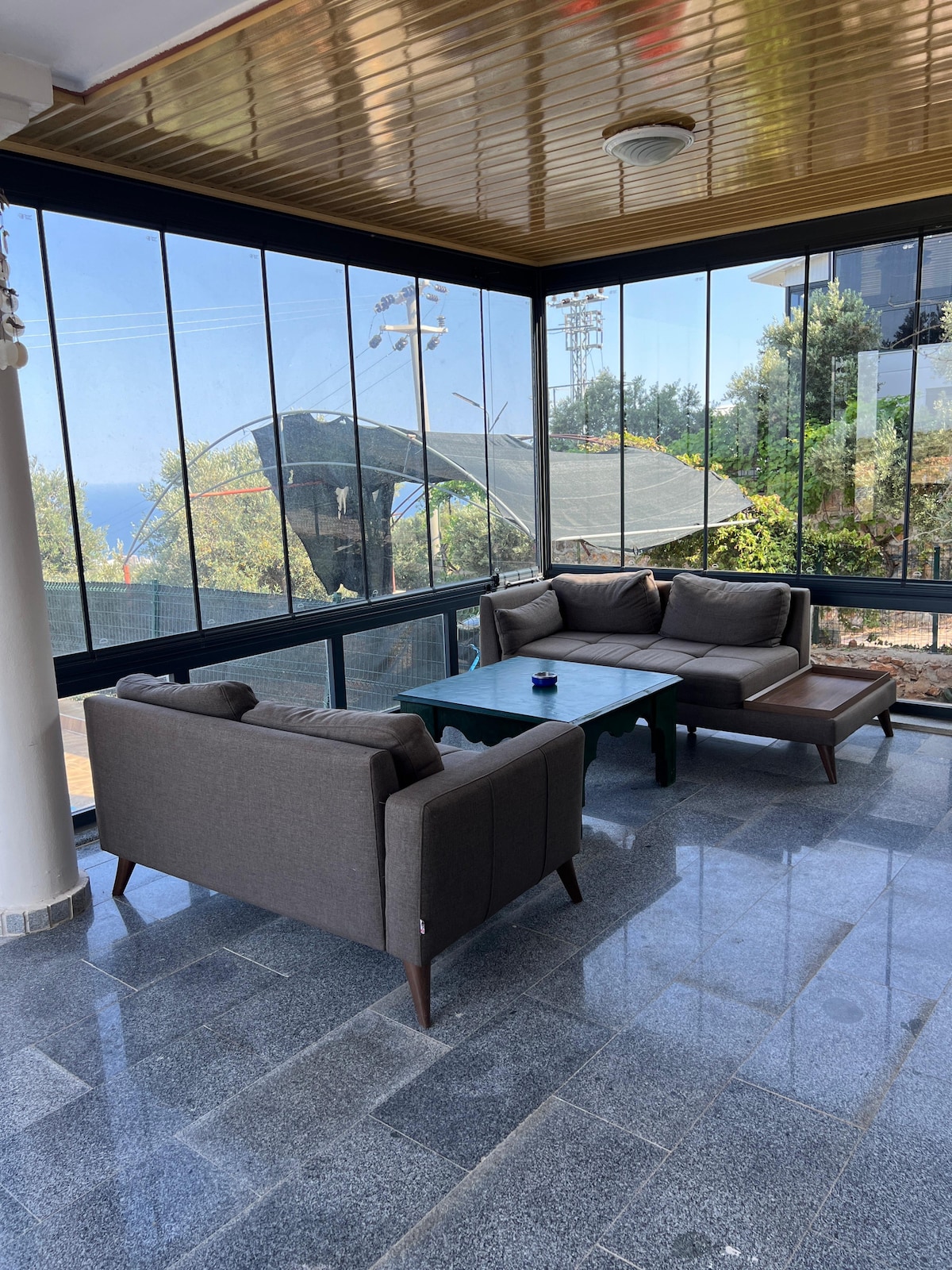 A sunlit seating area is featured with modern, comfortable furniture against a backdrop of large glass windows. The space includes a sofa set and a coffee table, with views of the surrounding greenery and the ocean visible outside.
