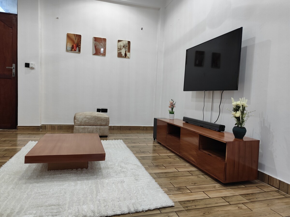 A living area features a low wooden coffee table and a light-colored rug, creating a cozy gathering space. A comfortable chair sits adjacent. The wall displays three framed art pieces, while a flat-screen TV is mounted above a wooden media console, enhancing the simplicity of the design.