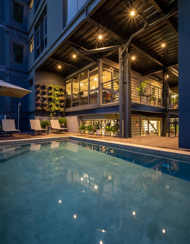 An inviting pool area features a serene swimming pool surrounded by lounge chairs. The contemporary building's façade is illuminated, showcasing expansive glass windows and greenery, creating a welcoming ambiance for relaxation and leisure.
