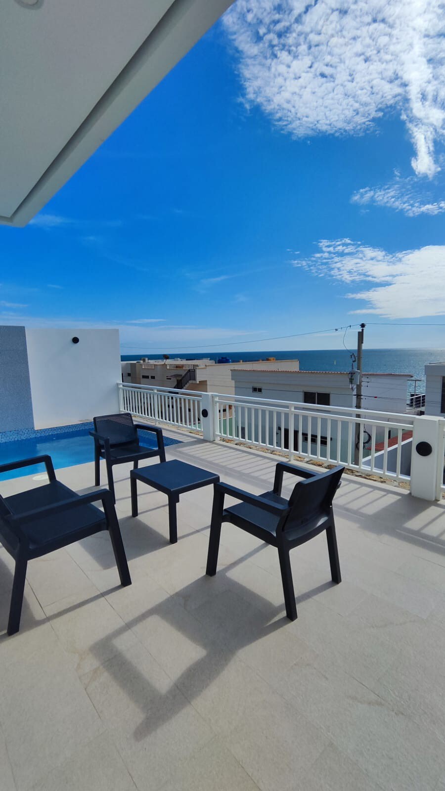 A spacious terrace overlooks the ocean, featuring four black chairs arranged around a small table. The view includes a clear sky and the distant coastline, complemented by a shimmering private pool at the edge of the area.