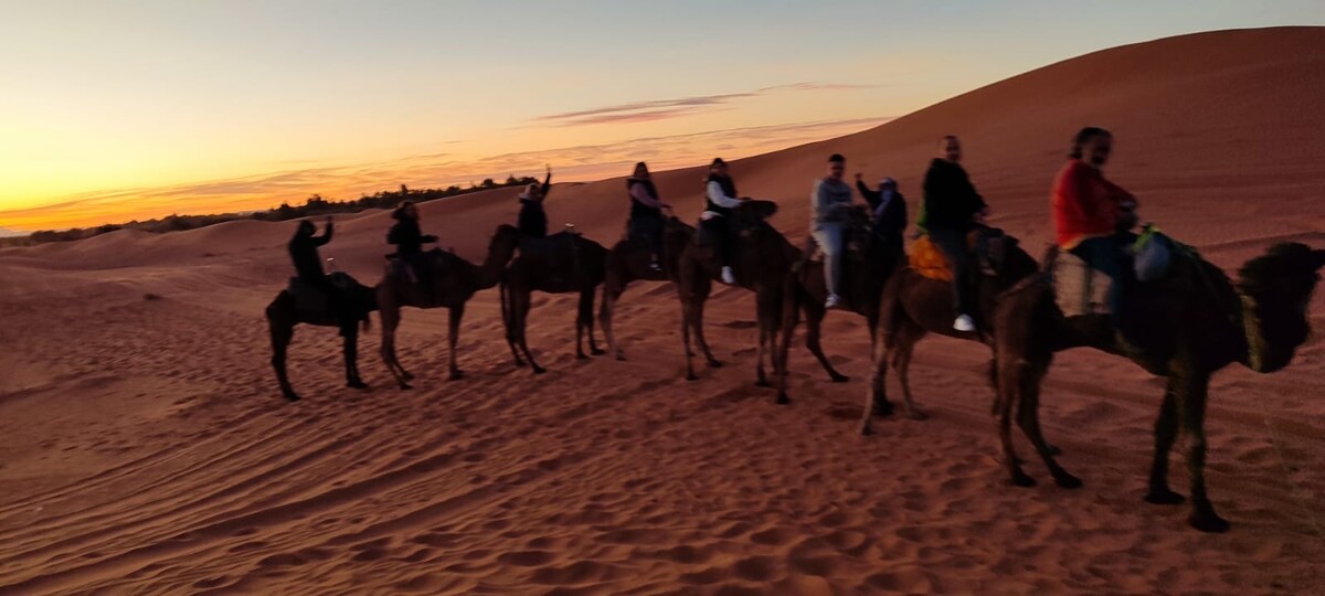 A line of camels is seen walking across the sand dunes at sunset, with riders seated atop each camel. The warm colors of the sky transition from orange to purple as the sun sets in the distance. The soft contours of the dunes create a serene backdrop.