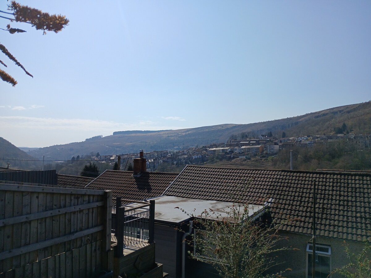 A scenic view is presented from the property, showcasing an expansive landscape of rolling hills and distant mountains under a clear blue sky. The rooftops of nearby houses are visible in the foreground, contributing to the picturesque outlook.