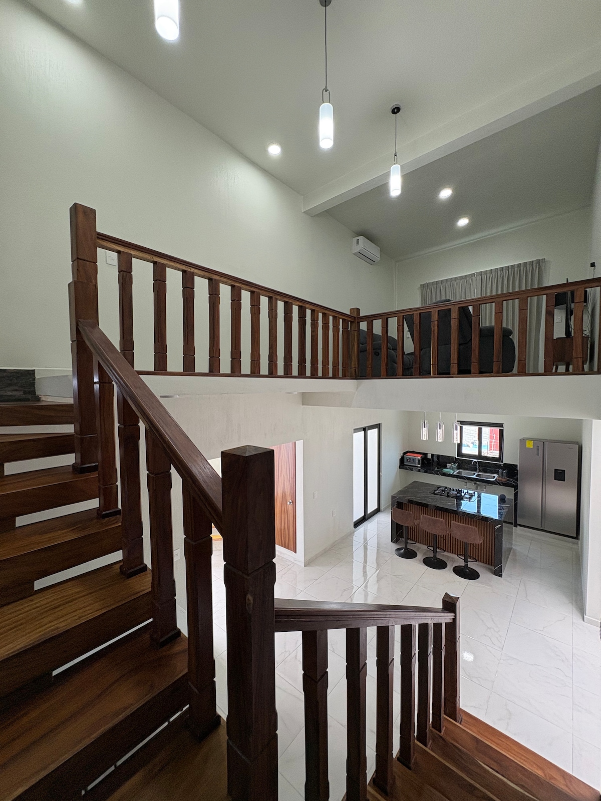 A spacious interior is showcased, featuring a wooden staircase leading to a second level. Bright lights hang from the ceiling, illuminating the open layout that includes a modern kitchen with bar stools. Large windows allow natural light to fill the area.