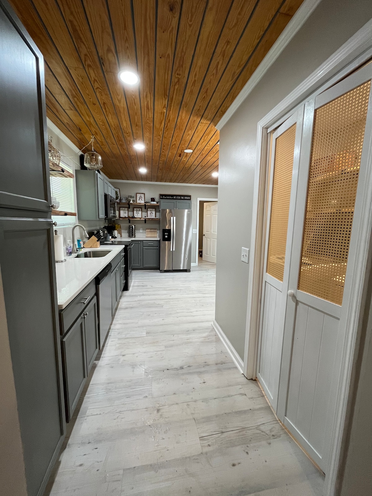 A modern kitchen corridor is highlighted by a wooden ceiling and neutral-toned cabinetry. A stainless steel refrigerator stands at the end, while the counter displays essential kitchen items. A door with a mesh panel leads to a pantry area, adding a functional touch to the space.