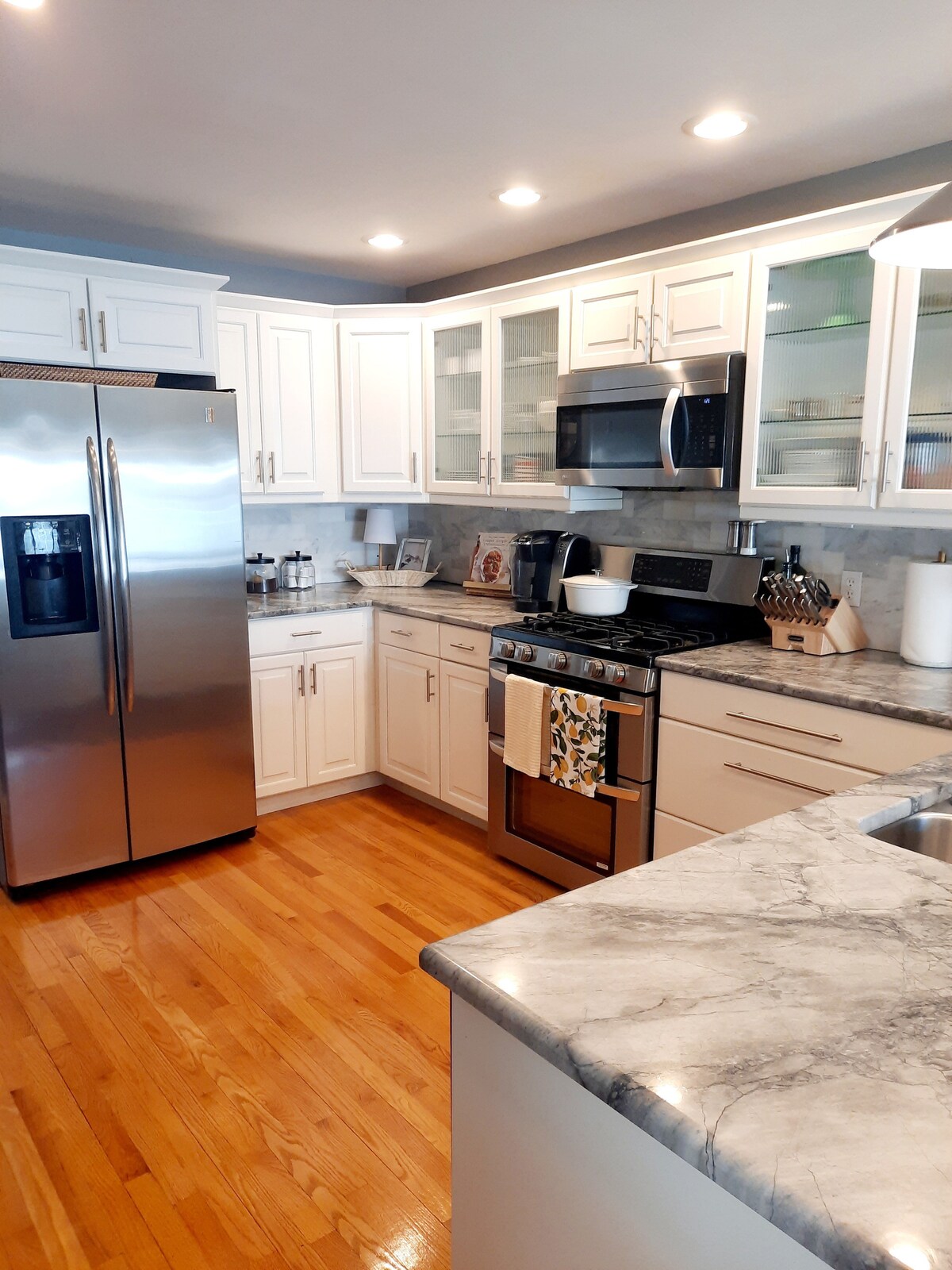 A modern kitchen is showcased with white cabinetry, contrasting dark marble countertops, and stainless steel appliances. Ample storage is provided by upper glass-front cabinets. A spacious refrigerator stands beside the sleek gas stove. Warm wooden flooring complements the overall aesthetic.