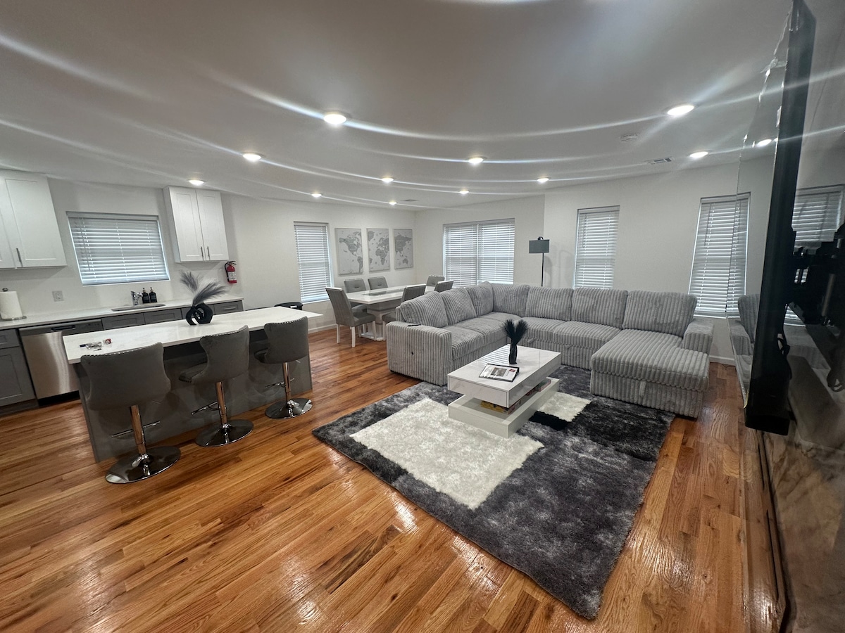 An open living space features a large sectional sofa arranged around a sleek coffee table. A soft area rug adds texture to the hardwood floor. The kitchen area displays modern bar stools and a stylish countertop, with ample lighting illuminating the room.