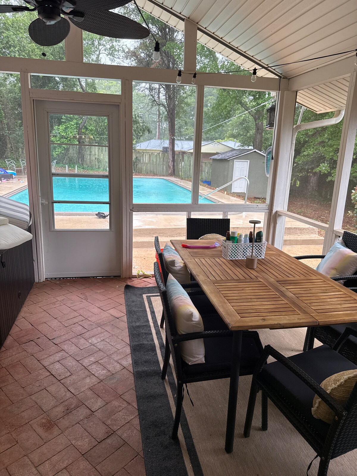 A screened-in porch features a wooden dining table surrounded by black chairs. A cozy rug is placed underneath. Beyond, a pool can be seen, with loungers positioned nearby. Lush greenery surrounds the area, enhancing the serene environment.