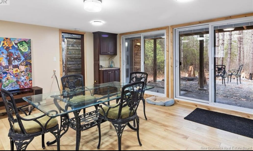 A dining area features a glass table surrounded by black metal chairs, accentuating the open space. Large sliding glass doors provide a view of the natural surroundings, while wooden flooring adds warmth to the room. A small kitchenette area is visible in the background.