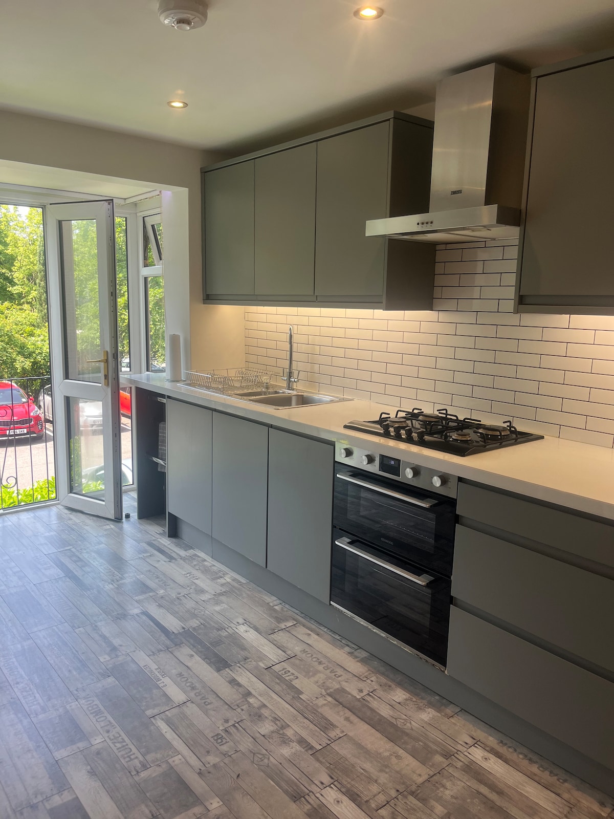 A modern kitchen is displayed with sleek gray cabinetry and a white tile backsplash. A gas stove and oven are positioned below a stainless steel hood. Natural light enters through an open door, illuminating the countertop and adding a spacious feel to the area.