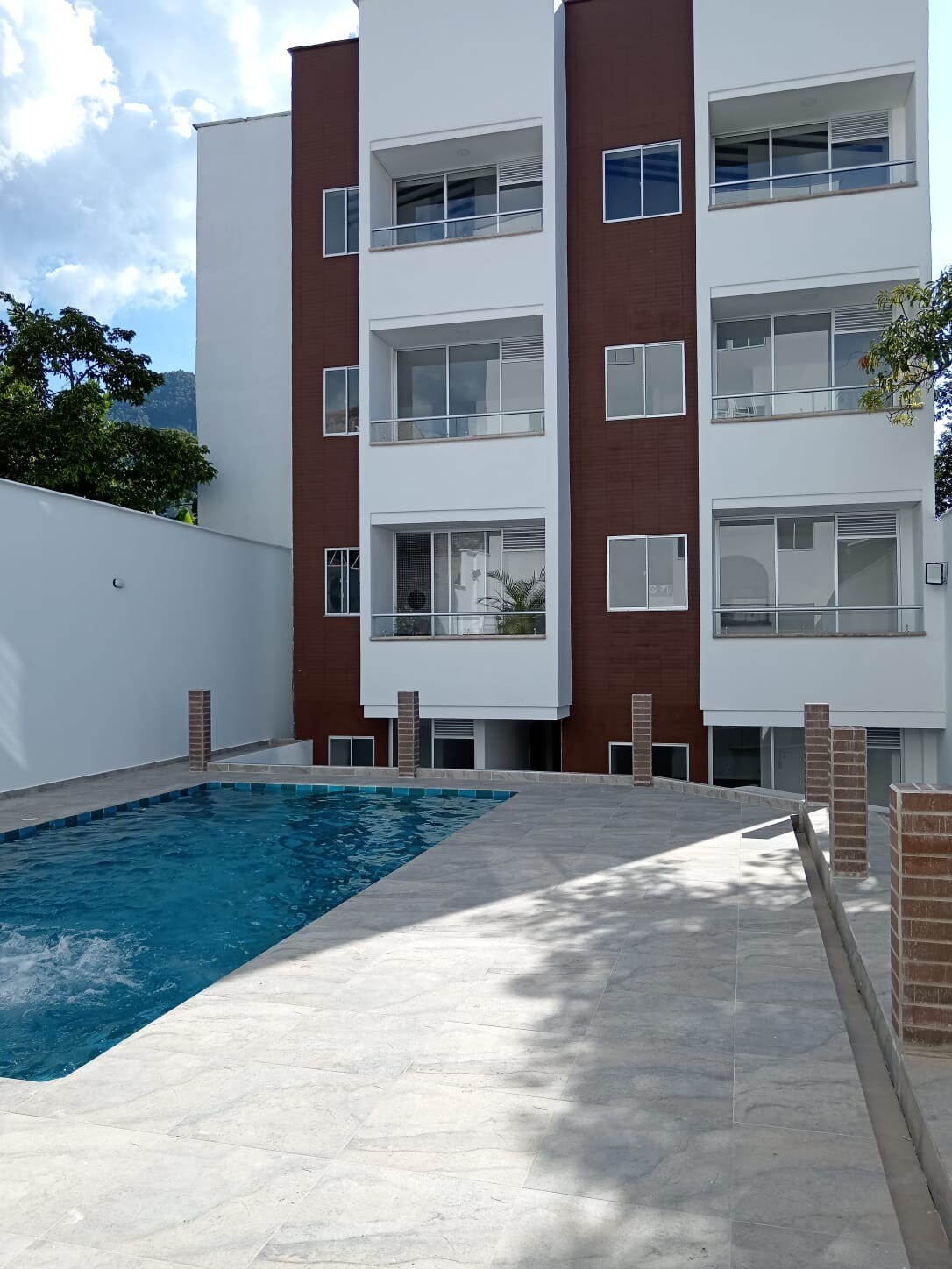 The exterior of a three-story building is featured, showcasing large windows and a modern design. A clear swimming pool, surrounded by a spacious stone patio, reflects sunlight, with green trees in the background enhancing the serene environment.