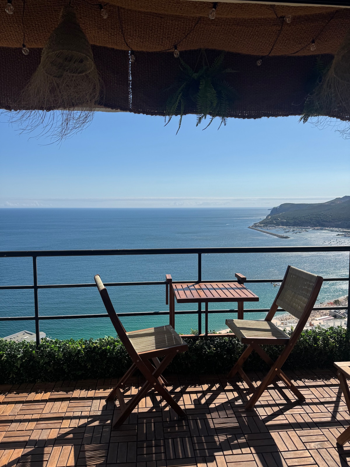 A private balcony is visible, featuring a small wooden table and two beige chairs. The ocean stretches out in the background, with a clear blue sky and gentle waves. The scene captures a serene view of the coastline, enhancing the overall tranquility.