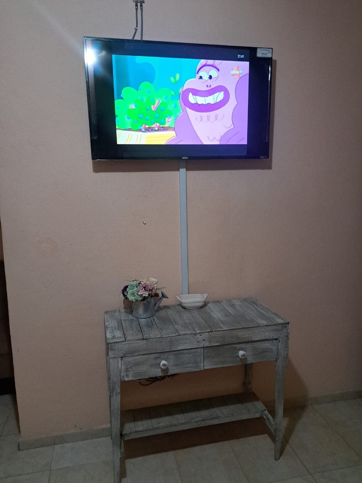 A wall-mounted television is displayed at eye level, presenting a colorful animated scene. Below, a rustic wooden table features a small flower arrangement and a decorative bowl, adding a simple touch to the room's design.