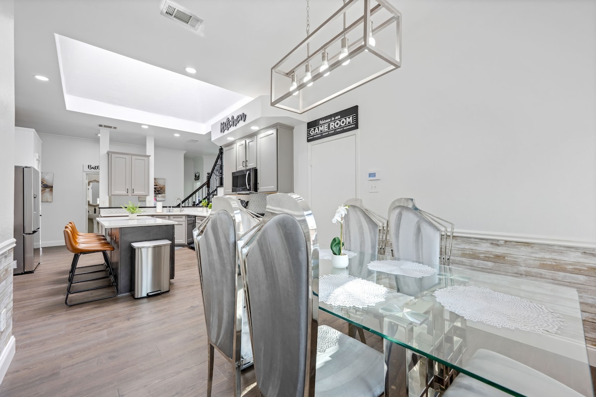 An open-concept dining area is presented, featuring a glass dining table surrounded by six elegant chairs. Natural light streams through the skylight overhead, illuminating the spacious kitchen that is visible in the background, equipped with modern appliances and a central island.