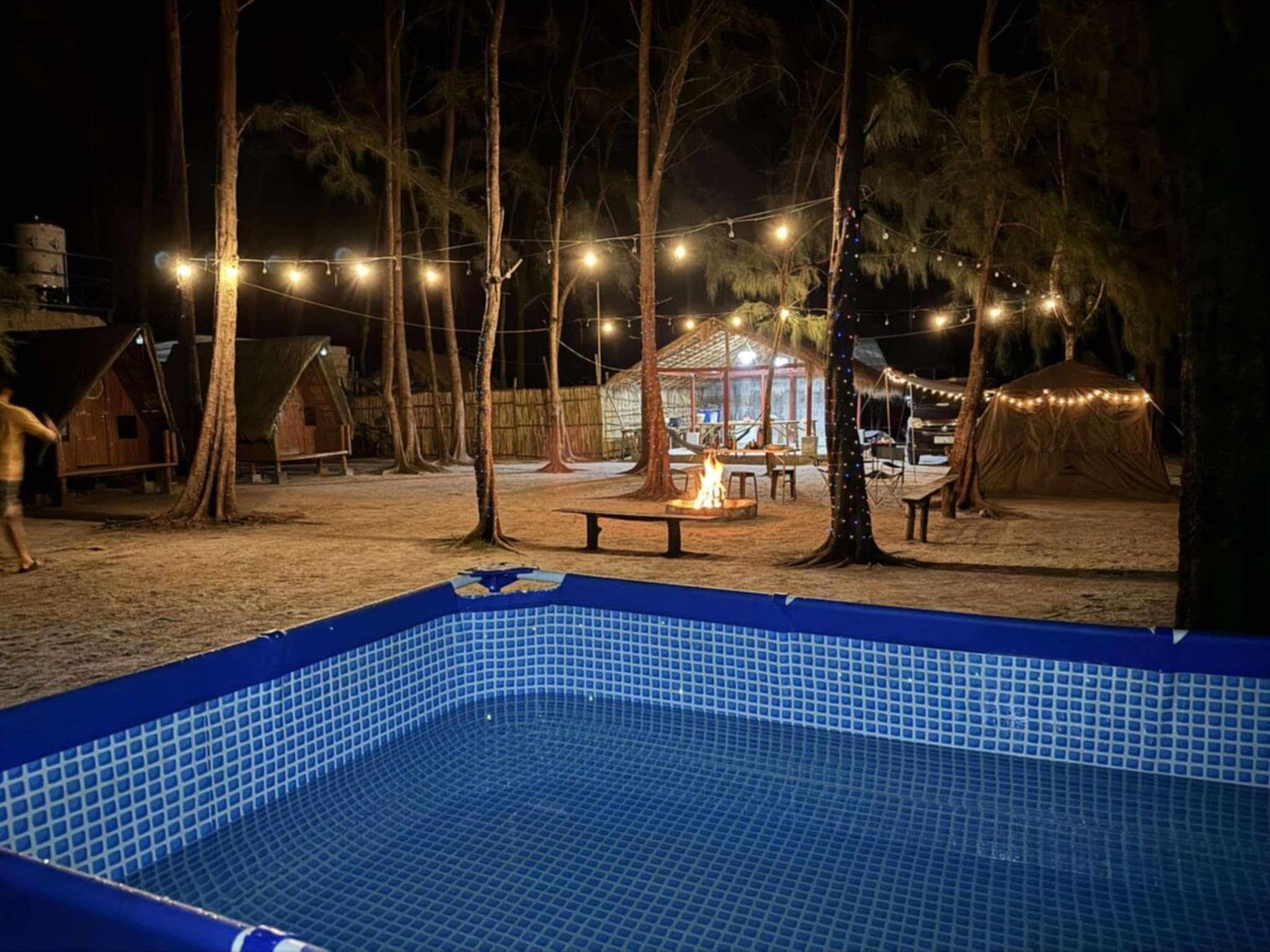 A swimming pool is visible at the forefront, surrounded by native huts and picnic tables. String lights illuminate the area, enhancing the ambiance of the outdoor space, while a bonfire area is positioned nearby among the trees, inviting evening gatherings.