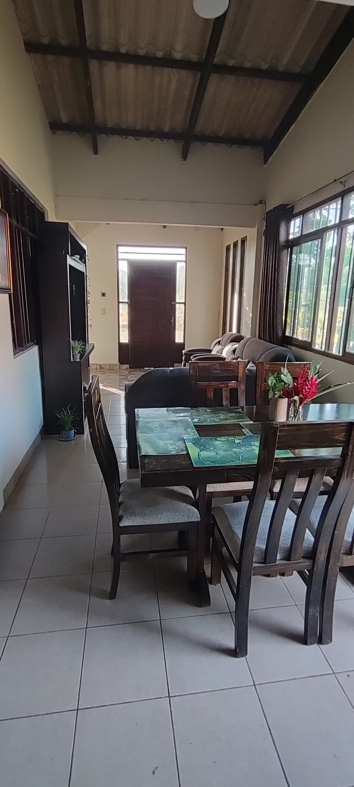 A dining area featuring a wooden table with four chairs sits adjacent to a cozy living space. Large windows allow natural light to illuminate the area, while a comfortable sofa is visible in the background. The walls are painted in neutral tones, enhancing a spacious feel.