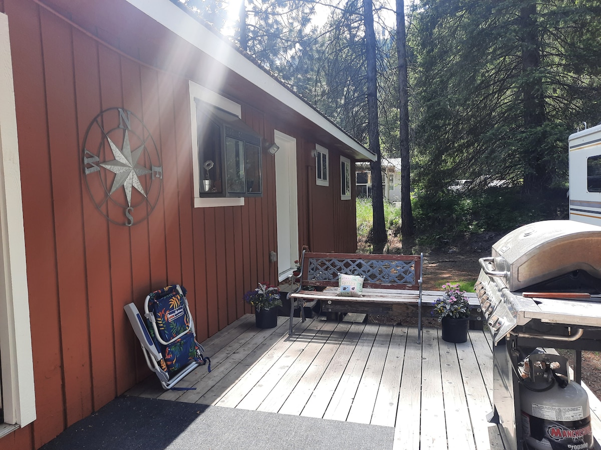A wooden deck extends alongside a rustic red cabin surrounded by trees. A wooden bench and potted plants enhance the outdoor space. A barbecue grill is positioned nearby, and a camping chair suggests a casual seating area. Sunlight filters through the trees, illuminating the setting.