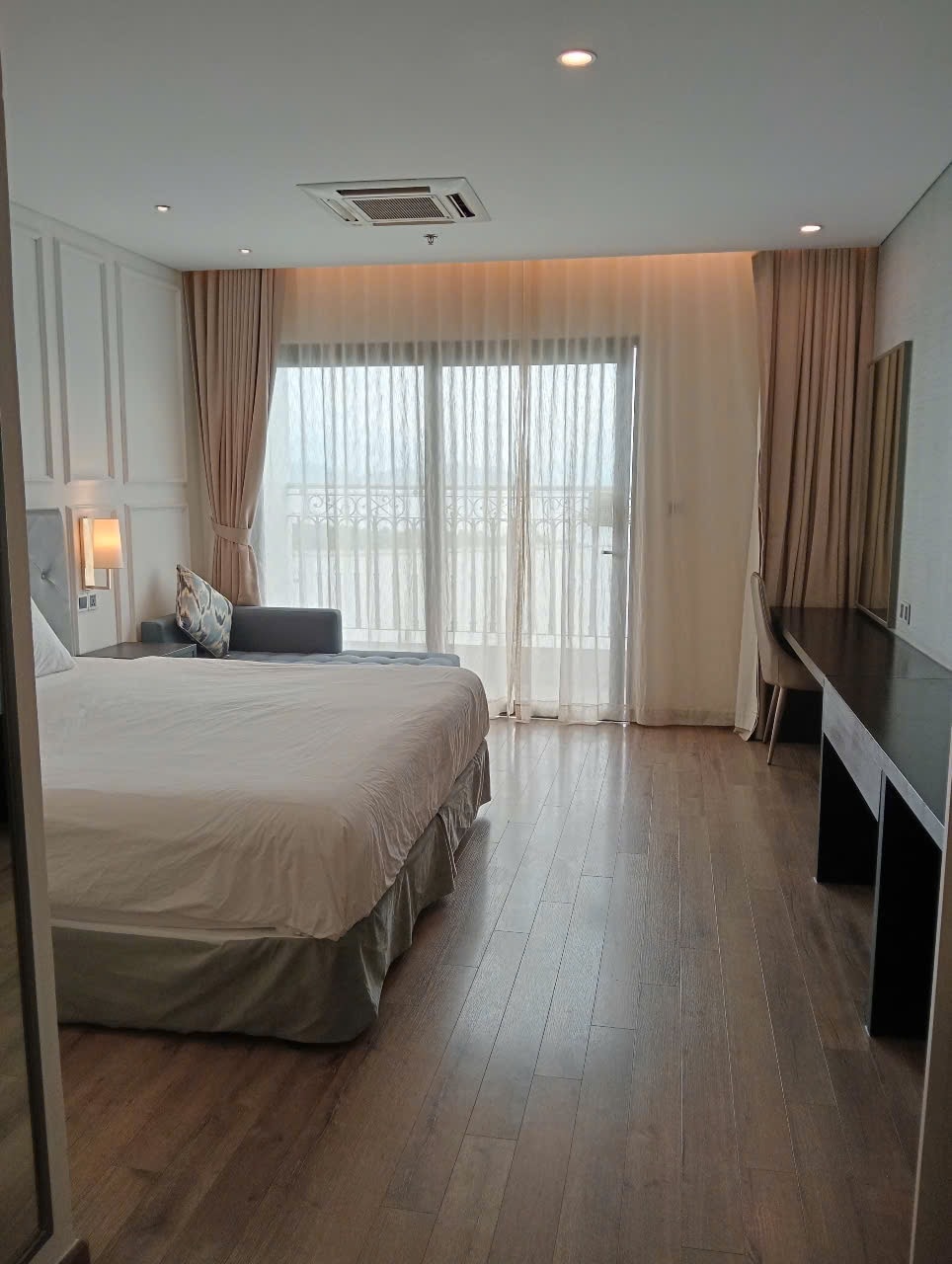 A bright and airy bedroom features a spacious bed with crisp white linens. Floor-to-ceiling windows provide natural light and unobstructed views, framed by sheer curtains. A comfortable chair and a sleek desk add functionality to the minimalist space.