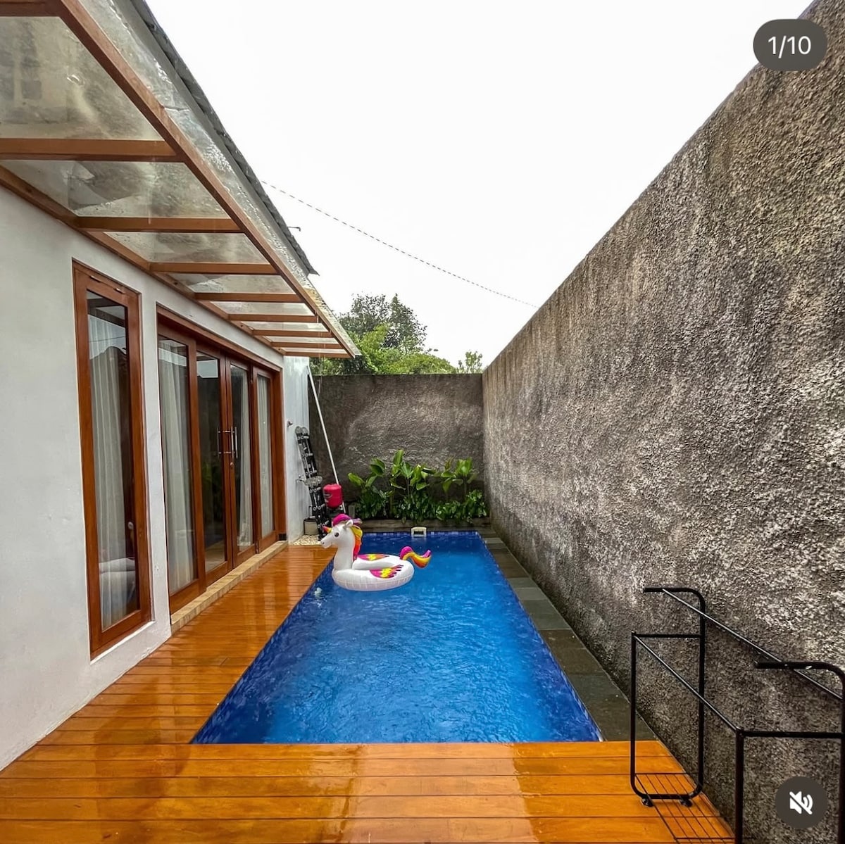 A refreshing swimming pool is framed by wooden decking and a smooth stone edge. A colorful inflatable pool float rests on the water's surface. The surrounding walls provide privacy, while greenery is visible beside the pool area. Large glass doors offer access to the indoor space.