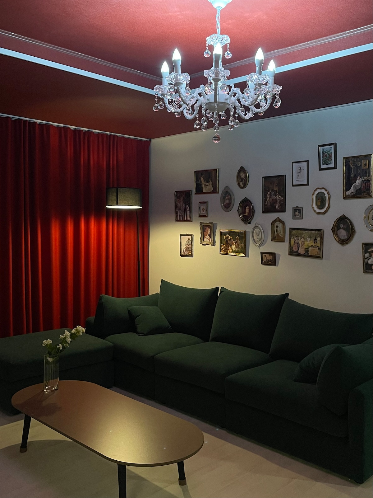 A cozy living area features a green sofa surrounded by various framed pictures on the walls. A small coffee table sits in front, while a chandelier provides ambient lighting. Red curtains frame the space, creating a warm and inviting atmosphere.