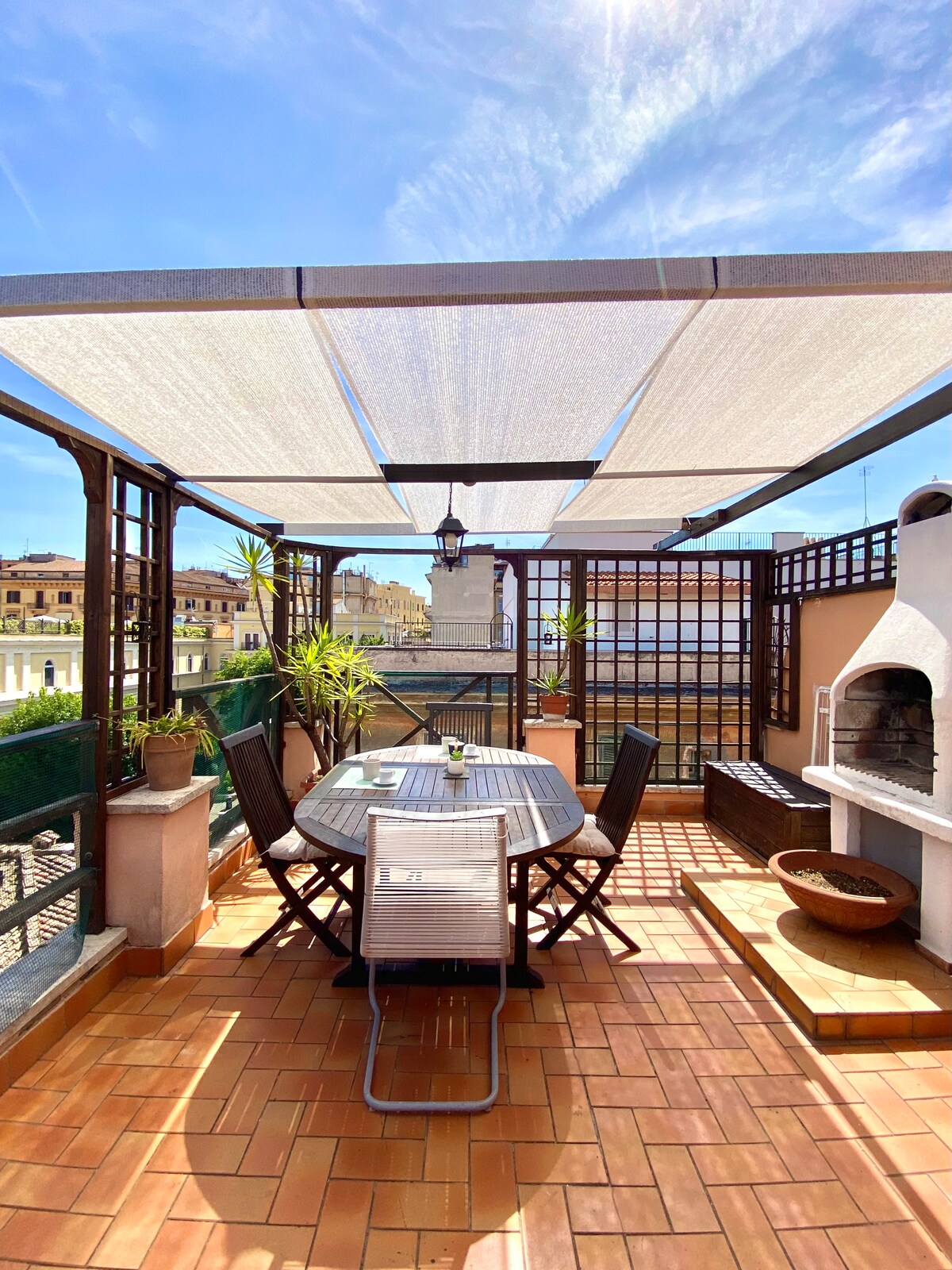 An expansive terrace is featured, equipped with a circular dining table and wooden chairs, shaded by a canopy. A brick barbecue is set against the wall, and surrounding greenery adds a touch of nature. Views of the historic buildings and rooftops of Borgo Pio are visible in the background.