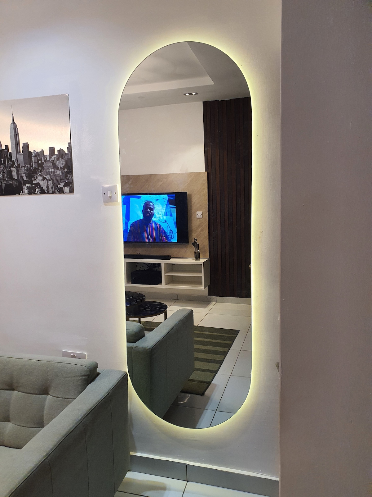 A large, elongated mirror with a soft glow is positioned against a wall. A contemporary living area can be seen through the reflection, featuring a light gray sofa and a TV stand with a television in view. The space is decorated with neutral tones and modern accents.
