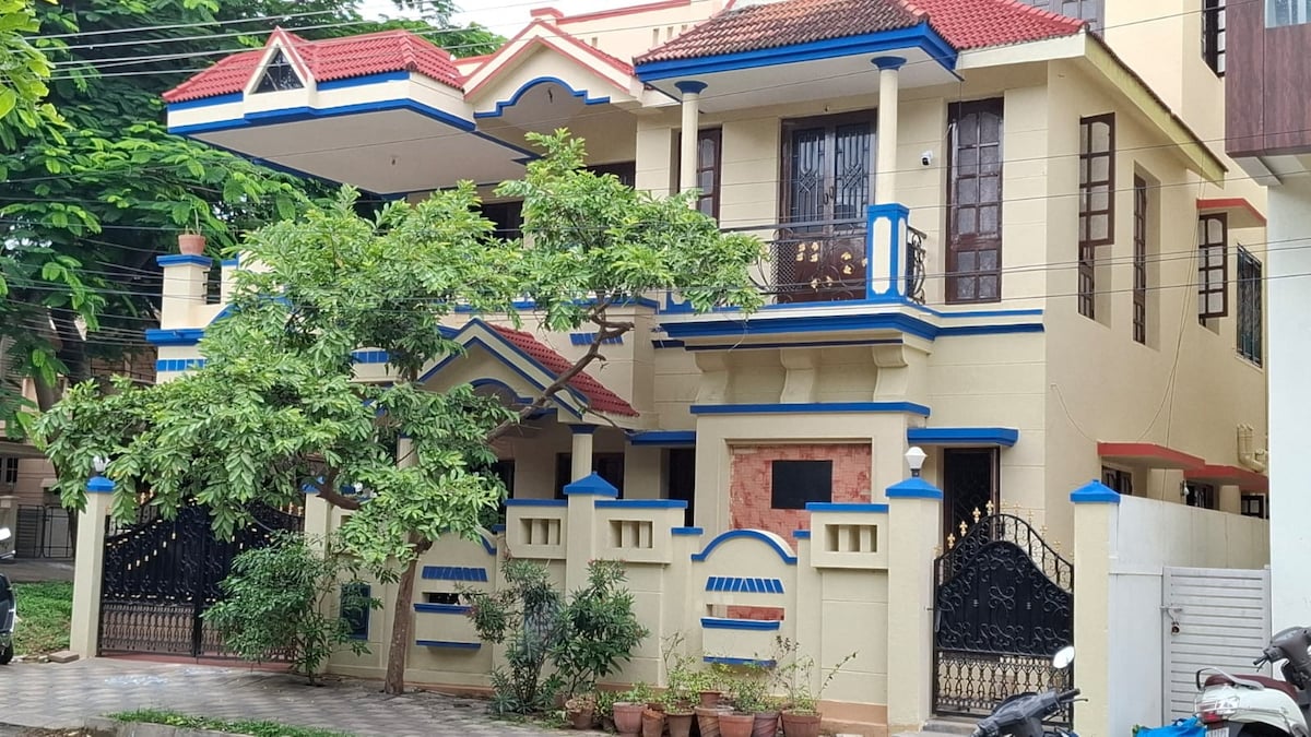 The exterior of the house is characterized by a combination of light-colored walls and blue accents. A prominent entrance with decorative features is framed by two gates. Lush greenery surrounding the property is complemented by the structural elements, including a balcony above.