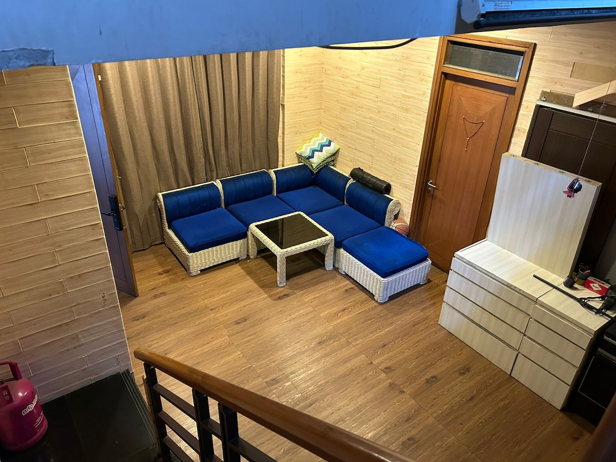 A spacious living area is furnished with a blue and white sectional sofa, complemented by a coffee table at the center. Soft, brown wooden flooring provides warmth, and a light-colored curtain covers the window, allowing natural light to fill the space. A doorway leads to another room.